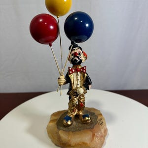 May include: A clown figurine standing on a textured amber-colored stone base. The clown wears a black and gold suit with a red and white polka-dot bow tie, holding three glossy balloons in yellow, red, and dark blue on thin metal wires.