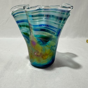 Pode incluir: Um vaso de vidro artesanal com uma forma orgânica única. O vaso apresenta faixas rodopiantes de azul, verde e turquesa, criando uma estética aquática vibrante. A borda tem um design ondulado, aumentando seu apelo artístico.