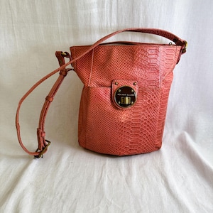 May include: A coral-colored bucket bag with a textured snakeskin pattern. The bag features a single adjustable shoulder strap and a circular brass-toned metal turn-lock closure on the front, which is engraved with the text Elliott Lucca.