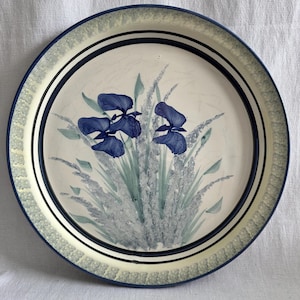 Puede incluir: Un plato redondo de cerámica con un diseño pintado a mano de flores de iris azul oscuro y follaje verde claro en el centro. El borde está decorado con una cenefa texturizada de color verde pálido y acabado con un filo azul oscuro y anillos negros.