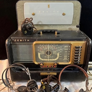 May include: Vintage Zenith Trans-Oceanic radio in a black case with gold accents. The radio has a dial, buttons, and the Zenith logo. Includes headphones and a speaker. The case is open, revealing the radio's interior.