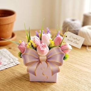 Pode incluir: Arranjo floral decorativo em uma caixa lavanda com um grande laço. O arranjo inclui tulipas rosa, flores amarelas e galhos verdes. Uma etiqueta diz "Crafted with Love".