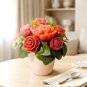 Könnte beinhalten: Ein Blumenarrangement mit geschnitzten Blumen in Rot-, Rosa- und Orangetönen mit grünen Blättern. Die Blumen befinden sich in einem hellrosa Topf. Das Arrangement steht auf einem runden Holztisch.