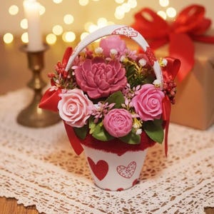 Könnte beinhalten: Ein dekoratives Blumenarrangement in einem weißen Korb mit Henkel. Der Korb ist mit roten Herzen verziert. Das Arrangement zeigt rosa und rote Rosen und andere Blumen. Eine brennende Kerze und ein Geschenk mit roter Schleife befinden sich im Hintergrund.