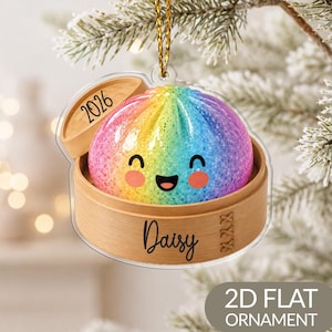 Könnte beinhalten: Ein flaches, transparentes Acryl-Ornament in Form eines Bambusdampfkorbs mit einem regenbogenfarbenen, glitzernden und lächelnden Teigtaschen-Charakter. Der Deckel zeigt das Jahr 2026 und der Boden ist mit dem Namen Daisy in schwarzer Schrift versehen.
