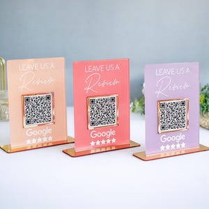 Peut inclure: Trois présentoirs de table en acrylique pêche, corail et lavande, chacun avec un code QR et le texte "LEAVE US A Review" et "Google". Les présentoirs ont une base dorée et sont exposés sur une surface blanche.