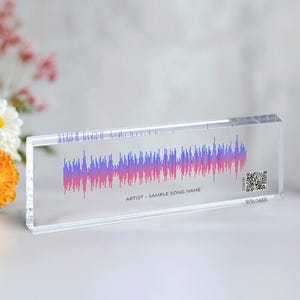 May include: A clear rectangular block featuring a pink and purple sound wave design. The text "ARTIST - SAMPLE SONG NAME" is printed below the sound wave. A QR code is visible on the right side of the block.