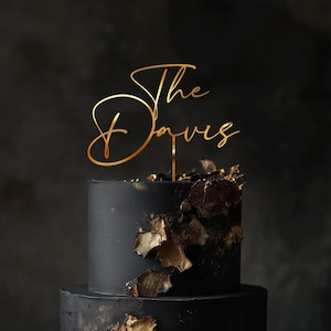 May include: A two-tiered black cake with gold leaf accents. A gold cake topper reads "The Davies" in elegant script. The cake is set against a dark background, creating a sophisticated look.