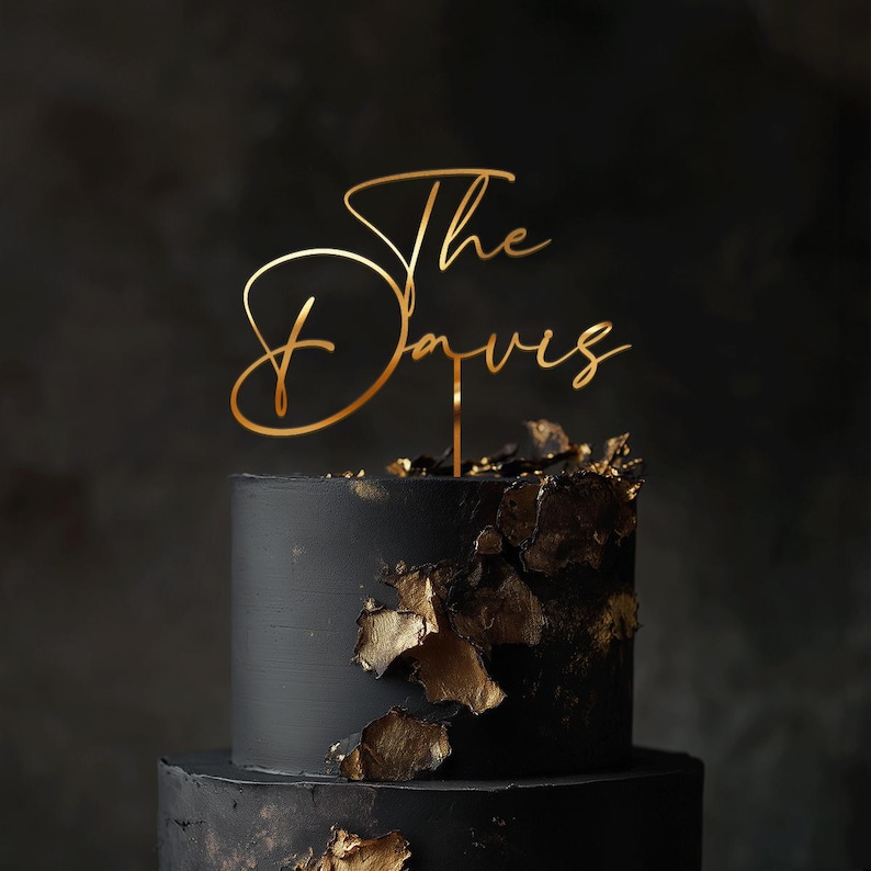 May include: A two-tiered black cake with gold leaf accents. A gold cake topper reads "The Davies" in elegant script. The cake is set against a dark background, creating a sophisticated look.