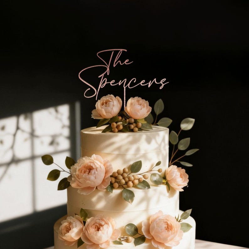 May include: A three-tiered wedding cake with peach-colored flowers and greenery. The cake is topped with a pink script cake topper that reads "The Spencers". The cake is decorated with peach-colored roses and small, round, golden berries.