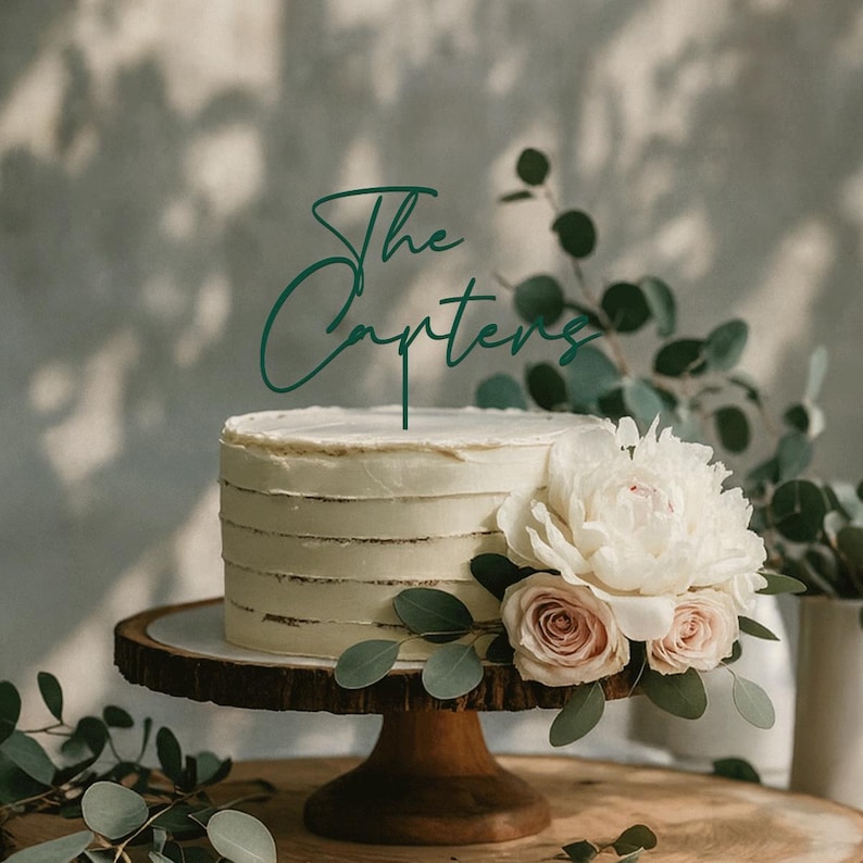 May include: A tiered white cake with a green topper that reads "The Carters." The cake is decorated with white frosting and sits on a wooden cake stand. The cake is adorned with white and pink flowers and greenery.