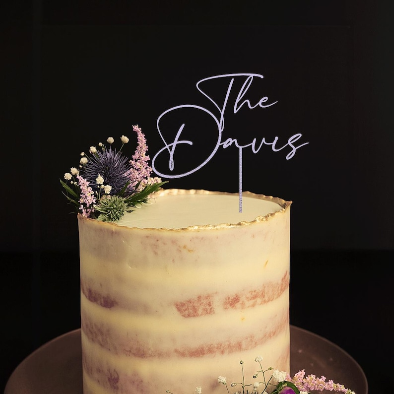 May include: A layered cake with a gold-trimmed top, adorned with a floral arrangement and a cake topper that reads "The Davies". The cake has a light-colored frosting and sits on a brown plate against a dark background.