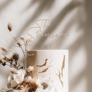 May include: A white wedding cake with gold accents and floral decorations. A wooden cake topper reads "The Spencers" in elegant script. The cake is adorned with white and tan flowers and greenery.
