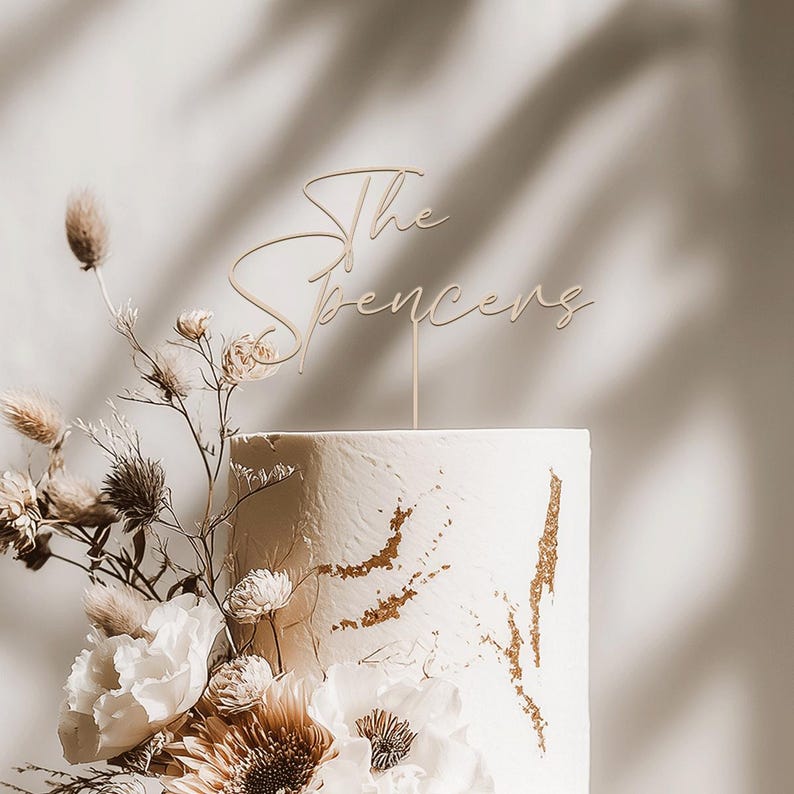 May include: A white wedding cake with gold accents and floral decorations. A wooden cake topper reads "The Spencers" in elegant script. The cake is adorned with white and tan flowers and greenery.