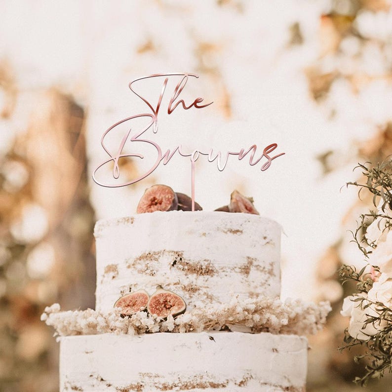 May include: A two-tiered white cake with fig slices and a decorative topper that reads "The Browns" in a rose gold script. The cake is decorated with dried flowers and greenery, perfect for a wedding or anniversary celebration.