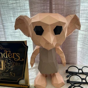 May include: A low-poly papercraft figure of a creature with large ears, a tan complexion, and black eyes, wearing a gray tunic. Beside it sits a black sign with gold text reading "Ollivanders" and "Fine Wands," along with black round glasses.