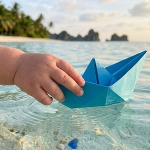 Może przedstawiać: Jasnoniebieska papierowa łódka origami unosząca się na czystej, płytkiej tropikalnej wodzie. Łódka ma klasyczny składany wzór ze spiczastym dziobem i centralną konstrukcją żagla, spoczywającą na powierzchni wody przy piaszczystym brzegu.