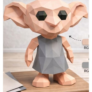 May include: A geometric papercraft figure of a creature with large ears, black eyes, and a gray dress. The figure is made of folded paper in shades of peach and gray, standing on a wooden surface.
