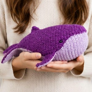 Peut inclure: Une peluche baleine crochetée avec un corps supérieur violet foncé et un ventre lavande clair. La baleine présente une tête arrondie, une petite nageoire dorsale, une nageoire caudale et un œil en perle noire sur le côté du corps en laine texturée.