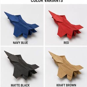 Op de afbeelding: Een raster van vier origami papieren vliegtuigen met het label COLOR VARIANTS. Links boven is marineblauw, rechts boven is rood, links onder is matzwart en rechts onder is kraftbruin. Elk straaljager-model heeft scherpe, geometrische vouwen.