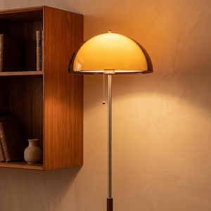 May include: A floor lamp featuring a dome-shaped amber glass shade and a slender metallic pole. The lamp includes a pull chain switch hanging from the center. The light emits a warm glow, illuminating the surrounding area with a soft radiance.