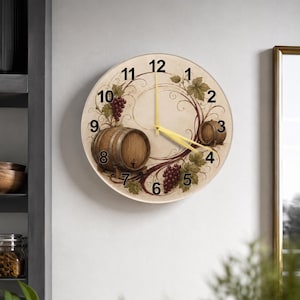 Puede incluir: Reloj de pared redondo con una esfera crema que presenta un diseño de barril de vino y uvas. El reloj tiene manecillas doradas y números negros. El diseño incluye ilustraciones de barriles de vino, uvas y hojas.