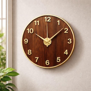 Puede incluir: Reloj de pared redondo con una esfera de madera marrón oscuro y manecillas y números dorados. El reloj tiene un borde dorado y está montado en una pared de color claro. Las manecillas del reloj indican que son aproximadamente las 10:07.