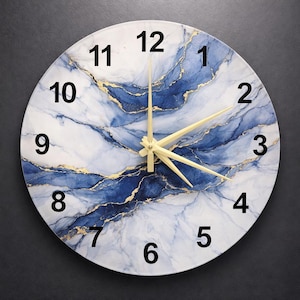 Puede incluir: Reloj de pared redondo con esfera blanca con diseño de mármol azul y dorado. El reloj tiene números negros y manecillas doradas. El diseño del reloj recuerda a la piedra natural.