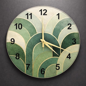 Op de afbeelding: Ronde wandklok met een groen en goud art deco ontwerp. De wijzerplaat heeft overlappende bogen in groentinten, met gouden accenten en zwarte cijfers. De wijzers van de klok zijn goud.