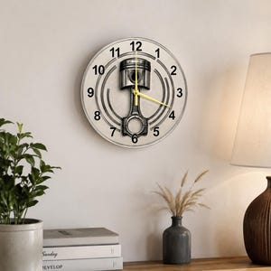 May include: A round wall clock featuring a central graphic of a metallic engine piston and connecting rod. The clock face is light gray with black numerals from 1 to 12 arranged in a circle. It has thin, bright yellow hour and minute hands.