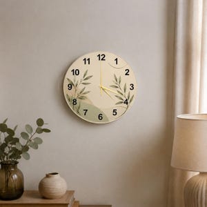 May include: A round wall clock with a cream-colored face featuring black numerals from 1 to 12. The design includes green botanical leaf illustrations and abstract shapes in muted tones. It has thin gold-colored hour, minute, and second hands.