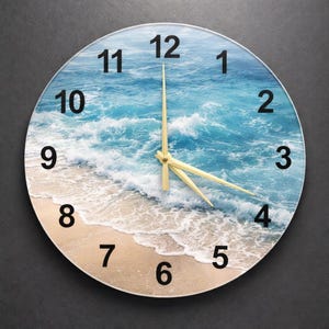 May include: Circular wall clock depicting a beach scene with blue ocean waves and a sandy shore. The clock face has black numerals and gold hands. A decorative item for home decor, with a diameter of approximately 30 cm.