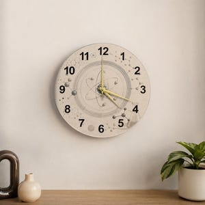 Puede incluir: Un reloj de pared redondo con un tema científico. La esfera del reloj presenta un diagrama de un átomo con números alrededor del borde. Las manecillas del reloj son amarillas. El reloj está montado en una pared de color claro.