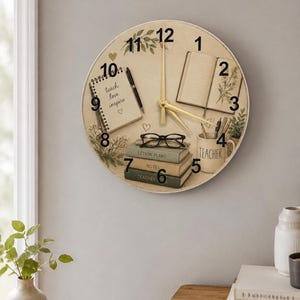 Könnte beinhalten: Eine runde Wanduhr mit beigem Hintergrund zum Thema Lehrer. Das Zifferblatt enthält einen Notizblock mit den Worten "teach love inspire", Bücher, eine Brille und eine Tasse mit der Aufschrift "teacher". Die Zeiger der Uhr sind goldfarben.