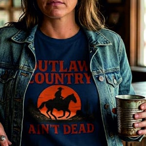 Puede incluir: Una mujer con sombrero de vaquero marrón, chaqueta vaquera y camiseta azul marino con el texto "OUTLAW COUNTRY AIN'T DEAD" y un gráfico de un vaquero a caballo. Sostiene una taza de metal.