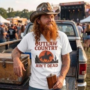 May include: White t-shirt with the text "OUTLAW COUNTRY AIN'T DEAD" in brown. The shirt features a graphic of a cowboy on a horse against a sunset backdrop. The person is wearing a cowboy hat and holding a brown flask.