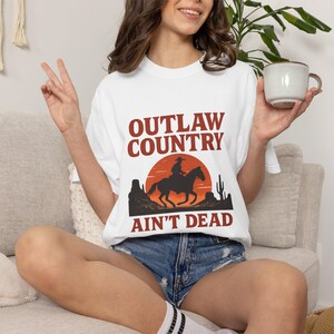May include: White t-shirt with the text "OUTLAW COUNTRY AIN'T DEAD" in brown and black. The shirt features a graphic of a cowboy on a horse in front of a sunset, with desert landscape silhouettes.