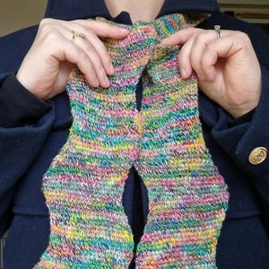 May include: A colorful, hand-knitted scarf with a vibrant mix of pink, yellow, green, and blue yarns. The scarf is held against a dark blue coat, showcasing its intricate knit pattern and bright hues. A unique accessory.