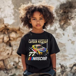 May include: Black t-shirt featuring a colorful NASCAR race car design with the number 77. The shirt includes the text "CARSON HOCEVAR" and "NASCAR" in white and yellow lettering. The shirt is worn by a young person.