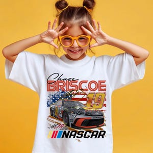 May include: White t-shirt featuring a graphic of a NASCAR race car with the text "Chase Briscoe 19" and "NASCAR". The design includes the American flag and racing flags. The shirt is worn by a person with orange glasses.