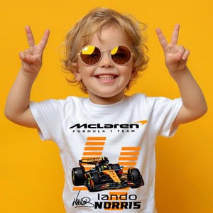 Könnte beinhalten: Ein weißes T-Shirt mit dem McLaren Formula 1 Team Logo und einer Rennwagen-Grafik. Das T-Shirt zeigt auch den Namen "Lando Norris". Ein lächelndes Kind mit runden Sonnenbrillen trägt das T-Shirt. Der Hintergrund ist gelb.