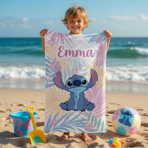 May include: A beach towel with a blue cartoon character design, set against a backdrop of pastel-coloured tropical leaves and flowers. The name "Emma" is printed in purple. A matching beach ball and sand toys are also visible.