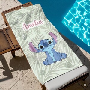 May include: A beach towel with a light green tropical leaf design and a cartoon character. The character is blue with large ears and eyes. The name "Amelia" is printed in pink. The towel is on a wooden sun lounger.