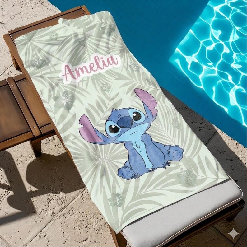 May include: A beach towel with a light green tropical leaf design and a cartoon character. The character is blue with large ears and eyes. The name "Amelia" is printed in pink. The towel is on a wooden sun lounger.