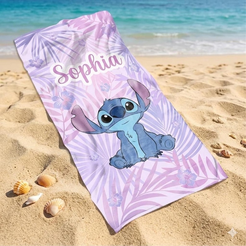 May include: A beach towel with a cartoon character in blue, with large eyes and ears, on a lavender background with a tropical leaf pattern. The name "Sophia" is printed on the towel.