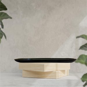 Può includere: Un vassoio rettangolare nero con un bordo leggermente rialzato, appoggiato su una pila di tre blocchi di legno colorati. Lo sfondo è un muro neutro e strutturato con piante verdi.
