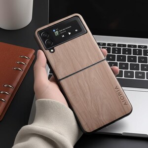 May include: A light brown phone case with a wood grain design, featuring the text "AIORIA" on the bottom. The phone displays a music player interface with the song "Over the Horizon". The case is open, revealing the phone's screen and camera.