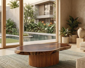 Op de afbeelding: Een ovale salontafel met een massief houten blad met natuurlijke rand in een donkerbruine afwerking en een cilindrische voet opgebouwd uit verticale houten latten. De tafel staat op een getextureerd vloerkleed in een kamer met grote glazen deuren.