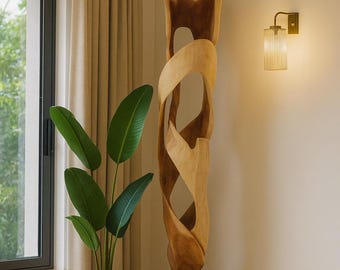 Op de afbeelding: Een hoge, abstracte houten sculptuur met een donkere basis staat naast een potplant met grote groene bladeren. Een wandlamp met een glazen kap zorgt voor warm licht. De kamer heeft een neutraal kleurenpalet.