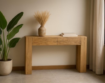 Op de afbeelding: Een lichte eikenhouten console met twee lades, bekroond met twee boeken en een decoratieve vaas met gedroogde bloemen. Een potplant met grote groene bladeren staat links. De kamer heeft een neutraal kleurenpalet.
